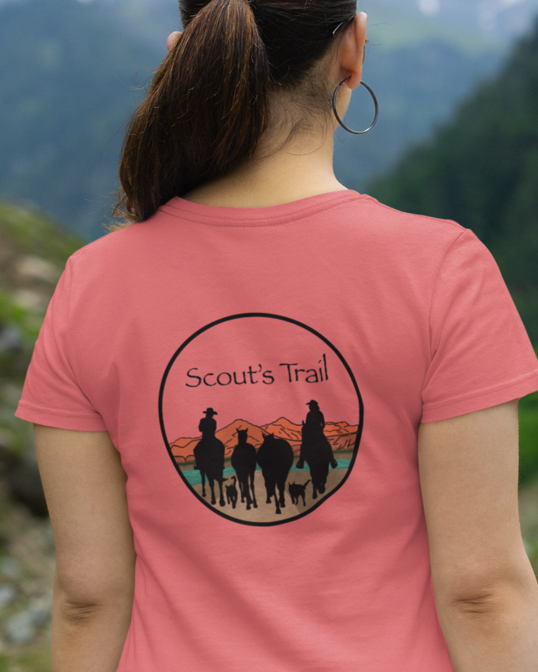 Scout's Trail logo. Silhouette of two riders with four horses and two trail dogs riding towards the mountains. Worn by a woman, logo on her back, looking out to mountains.