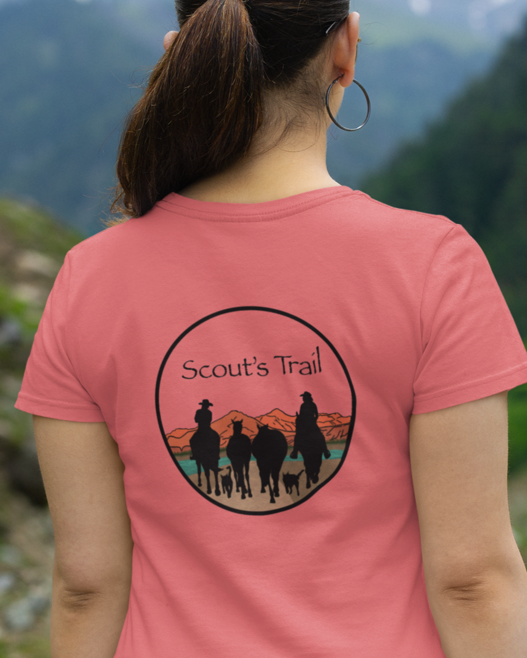 Scout's Trail logo. Silhouette of two riders with four horses and two trail dogs riding towards the mountains. Worn by a woman, logo on her back, looking out to mountains. 